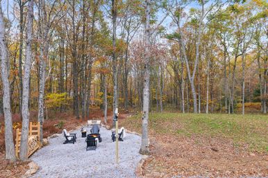 Cozy gravel backyard fire pit area with Adirondack chairs, stacked firewood and a lantern pole surrounded by tall trees and colorful autumn foliage in a wooded yard.