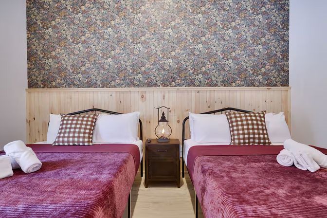 Cozy guest room with two burgundy velvet twin beds, checkered throw pillows, rolled white towels, central wooden nightstand with lantern lamp, rustic wood wainscoting and floral wallpaper.