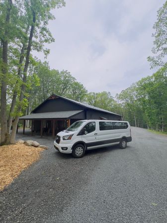 White passenger van parked on a gravel driveway beside a dark wood cabin in a green, forested rural retreat under an overcast sky
