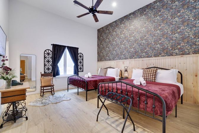 Cozy rustic guest room with two metal twin beds in burgundy throws, floral accent wall and wood wainscoting, ceiling fan, black-curtained window, vintage decor and hardwood floors — ideal for a countryside vacation rental.