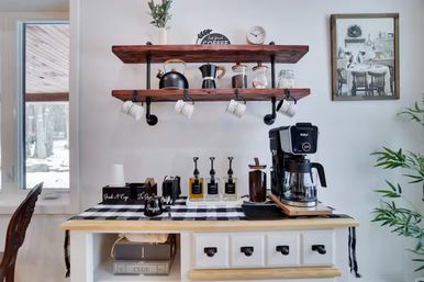 Cozy farmhouse-style kitchen coffee station with wooden pipe shelves holding mugs, kettle and jars, black-and-white buffalo check runner, syrup pumps, sugar jar and a drip coffee maker by a window with a snowy yard view.