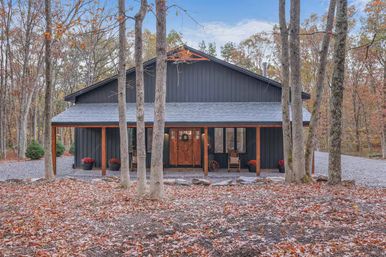 Cozy modern black cabin with wooden front door and covered porch in an autumn woodland, surrounded by tall trees, fallen leaves and a gravel driveway.