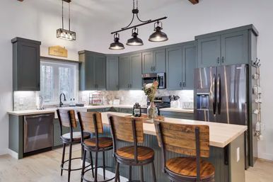 Modern farmhouse kitchen with green-gray cabinets, stainless steel appliances, butcher-block island with four wooden bar stools, subway-tile backsplash and industrial pendant lights
