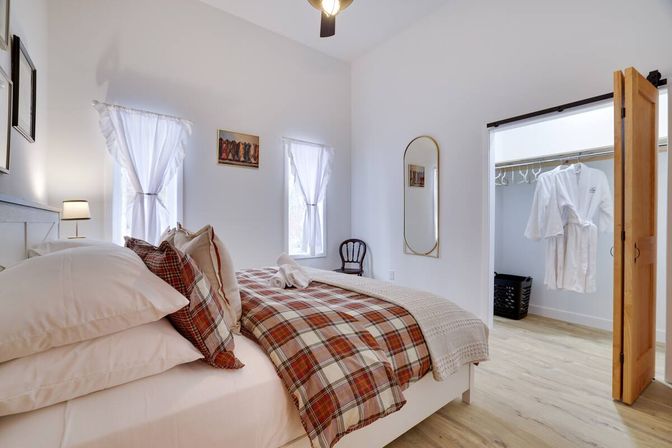 Cozy modern bedroom with plaid duvet and layered pillows, sheer-curtained windows, light wood floors, arched wall mirror and open walk-in closet with white robes.