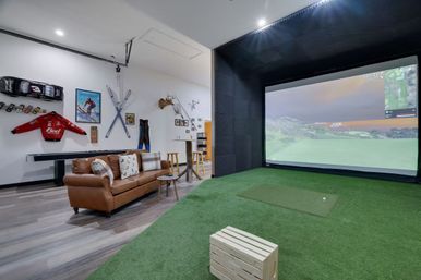 Home recreation room with indoor golf simulator — large projection screen over green turf and hitting mat, brown leather sofa with pillows, wooden crate and bar stools, wall-mounted skis, vintage racing jacket, model cars and antler decor.