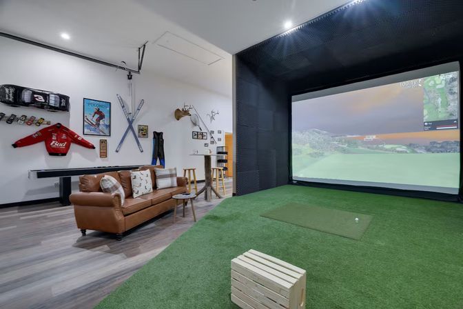 Cozy home recreation room with indoor golf simulator and large projection screen, artificial turf tee and golf mat, leather sofa, bar stools, skis, racing jacket and mounted deer heads.