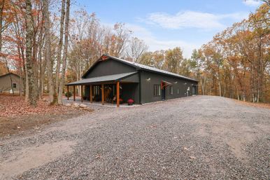 Modern dark-gray barn-style home with a covered wooden porch and long gravel driveway, nestled among tall trees showing orange and brown fall foliage in a rural wooded setting