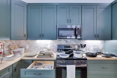 Modern residential kitchen with green shaker cabinets, stainless steel range and over-the-range microwave, light stone subway-tile backsplash, wooden countertops with pots, pans and bakeware, and an open utensil drawer.
