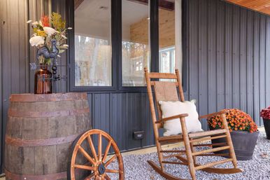 Cozy farmhouse porch with a wooden rocking chair and cushion, rustic barrel topped with a floral arrangement and rooster figurine, a leaning wagon wheel and potted orange mums against dark metal siding.