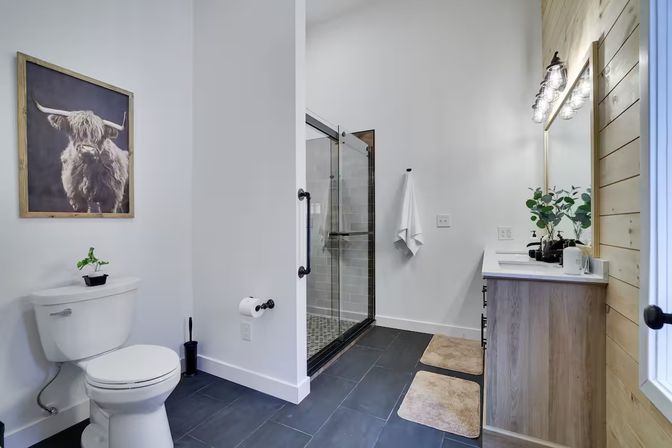 Modern farmhouse bathroom with black-framed glass shower, white walls, black tile floor, wood vanity and mirror with industrial lights, toilet with small plant and framed highland cow artwork, and beige bath mats.
