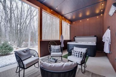 Cozy covered patio retreat with hot tub, four cushioned wicker chairs with check pillows, hanging robes and string lights overlooking a snowy woodland yard