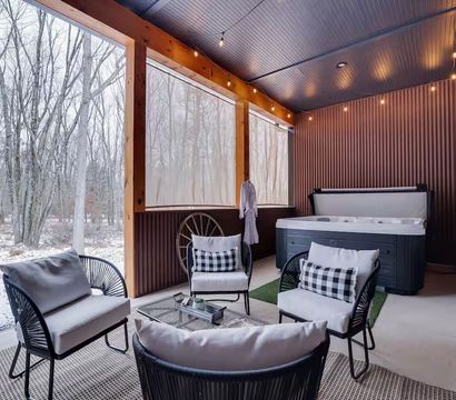 Covered rustic porch with hot tub, four cushioned lounge chairs and coffee table, string lights and hanging robes, overlooking snow-dusted woodland