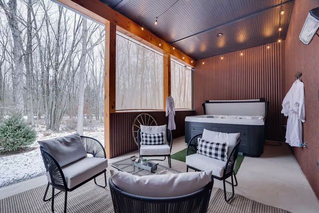 Covered rustic porch with hot tub, four cushioned lounge chairs and coffee table, string lights and hanging robes, overlooking snow-dusted woodland