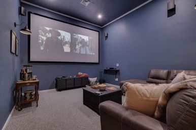 Cozy residential home theater media room with a large projector screen on deep-blue walls, brown leather sectional with pillows, central coffee table with snacks, side drink cart and carpeted floor — movie-night ready.