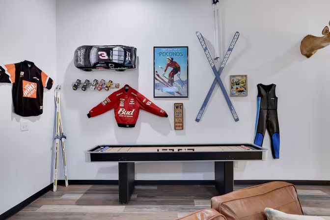 Bright modern basement game room with shuffleboard table, wall-mounted racing memorabilia (red racing jacket, model car, toy cars), crossed skis and vintage ski poster, wetsuit, mounted deer head, and leather sofa in the foreground.