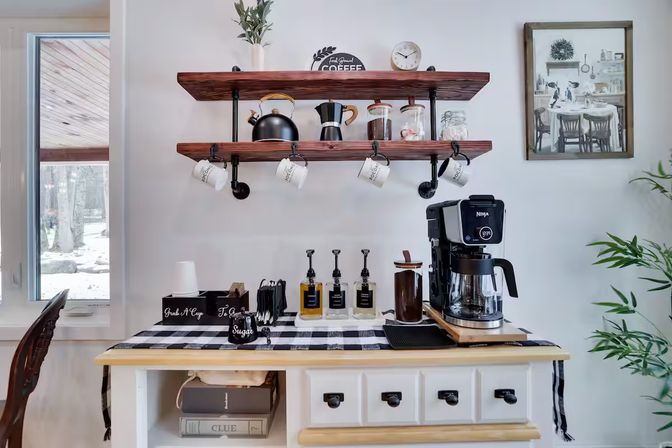 Cozy home kitchen coffee station with a black drip coffee maker on a white sideboard, wooden open shelves with hanging mugs, kettle, glass jars and syrup pumps on a black-and-white checkered runner — modern farmhouse style.