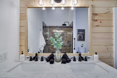Modern farmhouse double-sink bathroom with marble countertop, wood-framed mirror, black matte faucets, glass subway-tile shower and eucalyptus centerpiece
