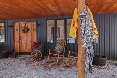 Rustic cabin covered porch with wooden ceiling and wreathed front door, two wooden rocking chairs by a barrel and wagon wheel, camouflage jacket and bright orange hunting vest hanging on a post, gravel floor scattered with fallen leaves.