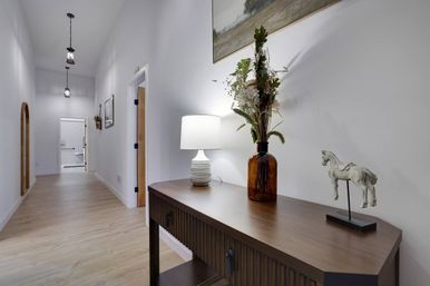 Bright minimalist hallway/entryway with light wood floors, wooden console table topped with a white lamp, amber vase of dried flowers and a small horse figurine, pendant lights and arched doorway in the background