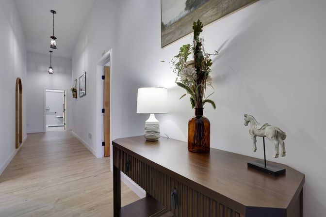 Bright modern home hallway entryway with light wood floors, white walls, hanging pendant lights, and a wooden console table topped with a lamp, amber vase of dried flowers, and a small horse sculpture.
