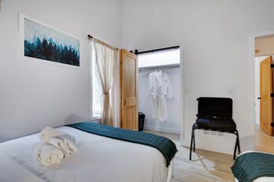 Bright minimalist guest bedroom in a vacation rental — white linens with teal runners, rolled towels on bed, open closet with hanging white robes, suitcase on luggage rack, light wood doors and framed forest print.