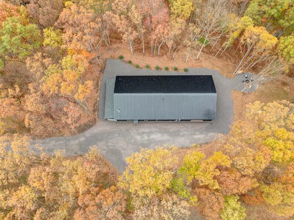 Aerial view of a long metal-roof building beside a gravel drive, nestled in colorful autumn woodland with vibrant orange, yellow and red fall foliage.