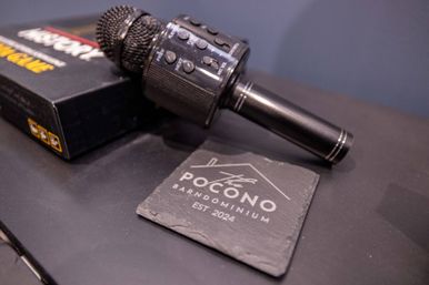 Black wireless karaoke microphone lying on a table next to a slate coaster and product box — close-up showing mic grille, control buttons and textured handle.