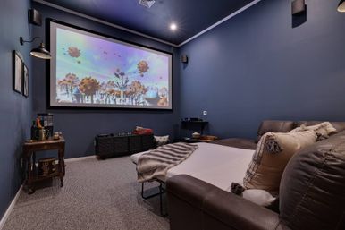 Cozy home-theater media room with large projector screen showing a colorful fantasy scene, navy-blue walls, wall-mounted speakers, carpeted floor, and a brown leather sectional pulled out with pillows and a faux-fur throw.