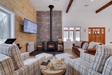 Cozy rustic cabin living room with plaid armchairs and ottomans facing a wood-burning stove on a stacked-stone hearth, wood-paneled wall with mounted TV, leather sofa, exposed beams and a snowy yard visible through the windows.