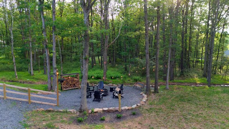 Cozy wooded backyard fire pit area with Adirondack chairs, stacked firewood, string lights and gravel patio beside a small creek