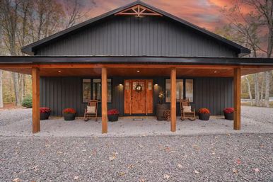 Modern black metal farmhouse with a wooden front porch and columns, rocking chairs and potted mums on a gravel driveway surrounded by autumn trees under a colorful sunset sky.