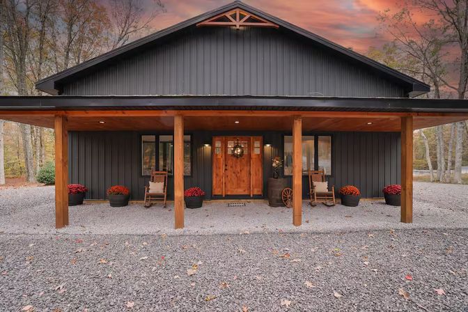 Cozy barn-style countryside home with dark metal siding, wooden covered porch, rocking chairs, potted mums and gravel driveway at autumn dusk.