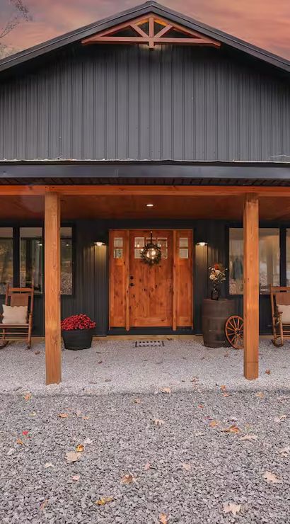 Cozy barn-style countryside home with dark metal siding, wooden covered porch, rocking chairs, potted mums and gravel driveway at autumn dusk.
