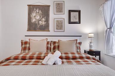 Cozy farmhouse-style guest bedroom with red plaid bedding, beige pillows, floral wall art, bedside lamp, and rolled white towels