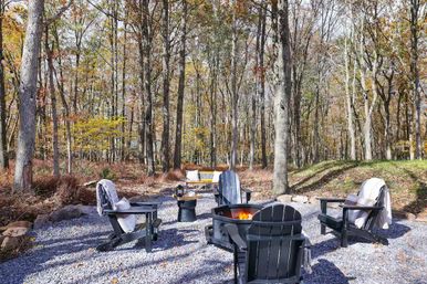 Cozy autumn woodland fire pit in a wooded backyard with black Adirondack chairs and throw blankets on a gravel seating area