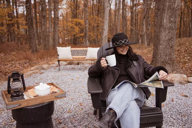 Woman in a wide-brim hat and shearling jacket sitting in an Adirondack chair on a gravel patio in an autumn forest, smiling while holding a mug and reading a magazine with a wooden bench, lantern and snack tray nearby.
