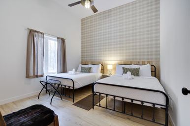 Cozy modern farmhouse guest room with two metal-framed double beds in white linens, plaid accent wall and wood headboard, bedside lamp and rolled towels, window with beige curtains, ceiling fan and light wood floors.