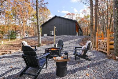 Cozy autumn cabin scene: four black Adirondack chairs around a glowing fire pit on gravel, stacked firewood and a modern dark cabin set in a wooded, fall-colored landscape.
