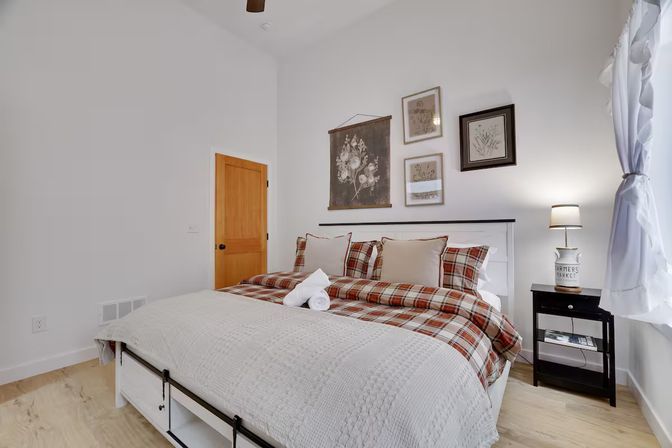 Cozy modern farmhouse bedroom with plaid duvet and white quilt, wooden door, framed botanical gallery wall, black nightstand with lamp, sheer white curtains and light hardwood floor.