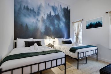 Modern guest bedroom with two metal‑frame beds in white linens and dark green throws, a large misty pine forest mural on the accent wall, bedside lamp, framed forest print and sheer‑curtained window letting in natural light.