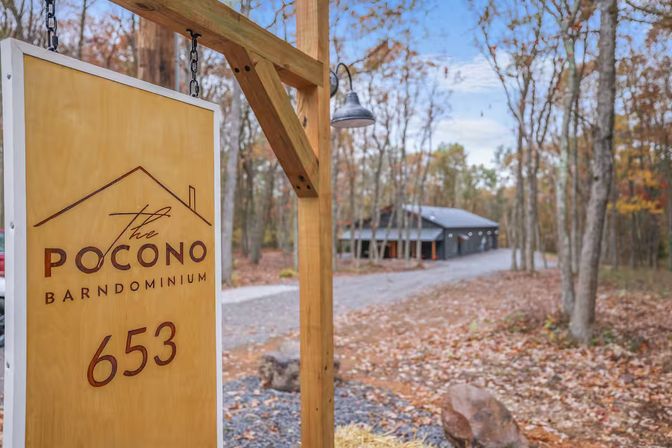 Wooden entrance sign hung from a timber post reading “653,” beside a gravel drive leading to a gray barn-style home surrounded by autumn woods.