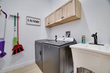 Bright home laundry room with matching dark metallic washer and dryer beneath natural wood wall cabinets, ‘LAUNDRY’ sign, hanging broom and mop, and white utility sink with black faucet and cleaning powder