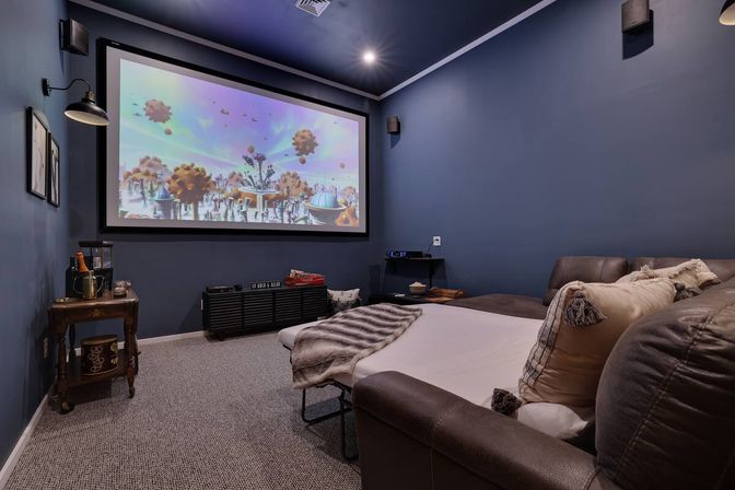 Cozy navy residential home theater with large wall-mounted projector screen showing colorful animation, leather sectional sleeper with pillows and blanket, carpeted floor and side table.