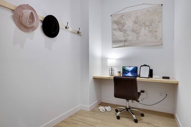 Cozy minimalist home office nook with a floating light-wood desk holding a laptop, lamp, small plant and mirror, a brown swivel chair, world map wall art, two hats on hooks and slippers on a light wood floor.