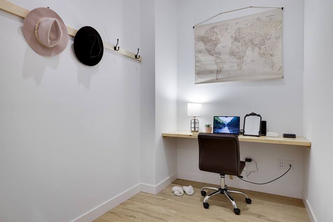 Cozy minimalist home office nook with a floating light-wood desk holding a laptop, lamp, small plant and mirror, a brown swivel chair, world map wall art, two hats on hooks and slippers on a light wood floor.