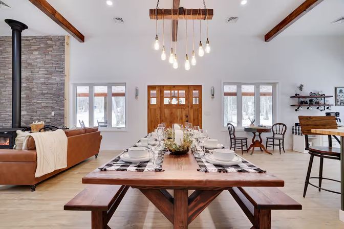 Open-concept farmhouse dining room with a long rustic wooden table set for dinner, Edison-bulb chandelier, leather sofa, stone wood-burning stove, and wooden front door