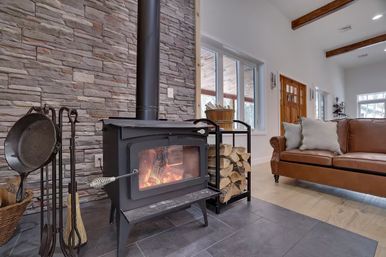 Cozy living room with black wood-burning stove and roaring fire against a stacked stone accent wall, firewood rack and tools, large windows, exposed wood beams, and brown leather sofa.