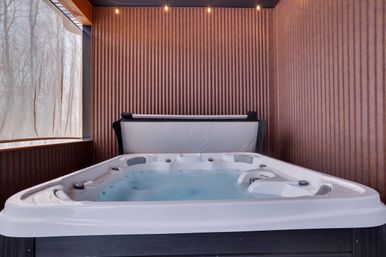 Cozy indoor hot tub in a wood-paneled private spa nook, bubbling water with folded cover, string lights overhead and a large window showing bare winter trees.