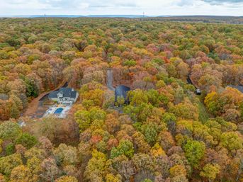 Aerial view of a private home with a backyard pool tucked into a densely wooded landscape of autumn foliage, vibrant orange, red, yellow and green trees stretching to the horizon with winding driveways visible.