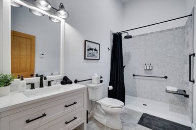 Contemporary white bathroom with shaker-style vanity, large mirror and industrial black lights, matte-black faucets and hardware, toilet, and accessible walk-in shower with black curtain, rain showerhead, gray tile, grab bars, and wall-mounted soap dispensers.